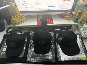 Three black baseball caps featuring a green logo, displayed in their packaging on a desk, with a small golden Buddha statue and other items visible in the background.