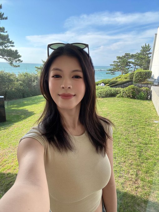 A selfie of a young woman with long dark hair wearing a beige top and sunglasses on her head, standing on a grassy lawn with the ocean and blue sky in the background.