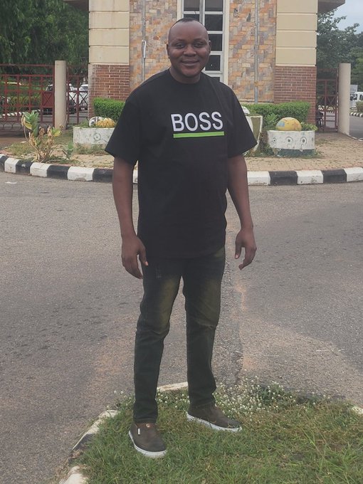 A man wearing a black t-shirt with 'BOSS' printed in white letters stands smiling on a paved area in front of a building.