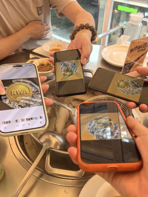 The image shows four cell phones being held up for viewing at an indoor restaurant table. The phones are displaying images of a gold coin and a spoon. The people around the table appear to be engaged with their phones, possibly sharing interesting content or discussing something related to the displayed items on their devices.