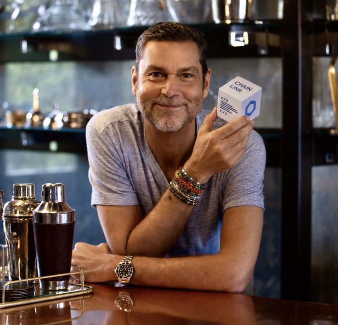 A man wearing a gray t-shirt and beaded bracelets smiles while holding a white cube labeled 'CHAIN LINK' next to his face.
