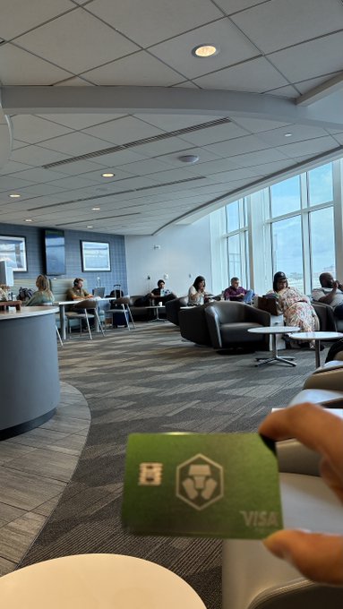 Inside an airport lounge, a hand holds up a green Visa credit card with a lion logo in the foreground, while passengers relax in seating areas near large windows in the background.