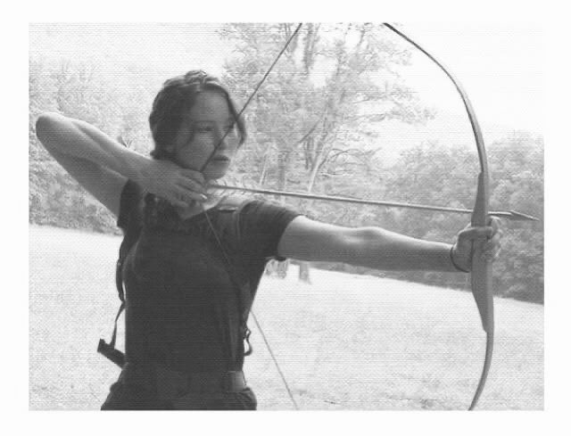 A young woman with dark hair, wearing a black t-shirt, is shown in a black-and-white photograph drawing back a bow and arrow, aiming with focused concentration in an outdoor wooded setting.