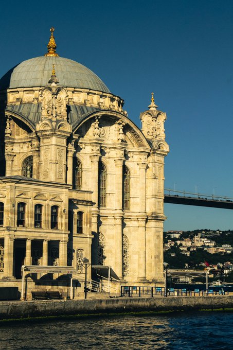 A grand domed stone building, likely a mosque, illuminated by sunlight against a clear blue sky, with a bridge visible in the background.