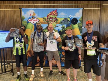 Five men wearing medals stand together indoors, holding what appear to be race medals, with a colorful inflatable play structure in the background.