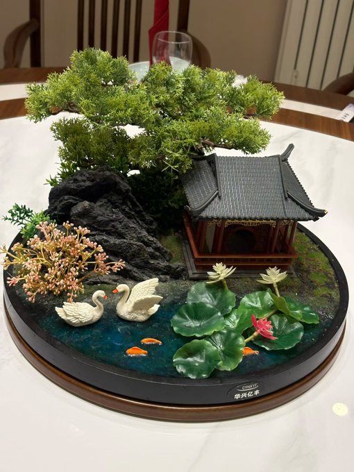 urn with a miniature garden scene, featuring a black roofed pagoda and two swans, surrounded by pink flowers and green plants on a white tablecloth.