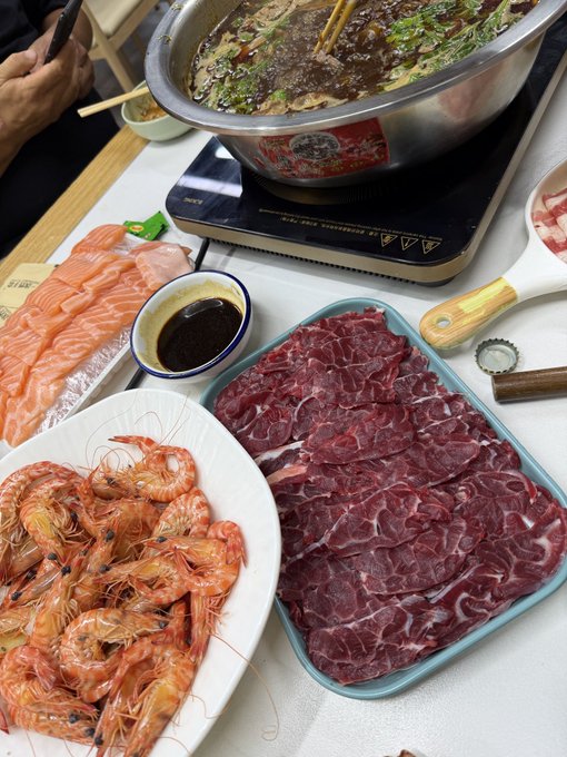 A spread of Chinese hot pot ingredients on a table, including a tray of thinly sliced raw beef, a plate of whole shrimp, sliced salmon, and a simmering pot of spicy broth on an induction cooker.