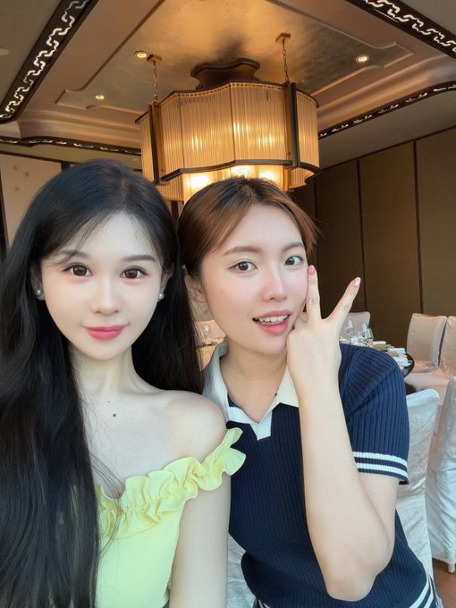 The image features two young women posing together for a photo, both wearing dresses. One of them has long black hair while the other has short brown hair. They are standing close to each other with their arms around one another's shoulders, creating an intimate atmosphere in front of a dining table and chairs. The background includes a chandelier hanging from the ceiling, adding elegance to the scene. The image also contains text that reads "遇见了又善良又温暖的Alina呀 @pearbinance 币圈的女子真的都好好呀 好多细节上啦".