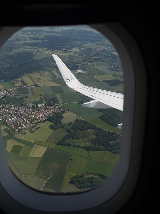 A view from an airplane window shows a Lufthansa wing extending over a patchwork of green farmland and small towns below, capturing the travel vibe of "Gm CT Beautiful Wednesday."
