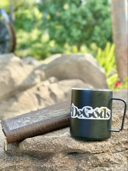 A black enamel mug reading 'DeGods' sits on a stone beside a worn Holy Bible, with green foliage in the background.