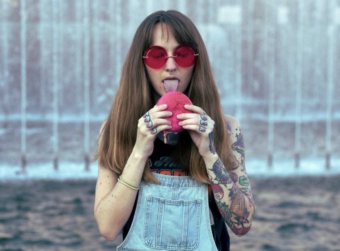 A young woman with long brown hair, red round sunglasses, and colorful tattoos on her arms wearing denim overalls, holding a pink ball with her tongue sticking out against a shimmering curtain backdrop.