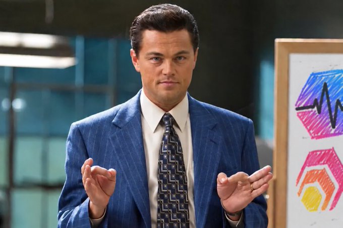 Leonardo DiCaprio, dressed in a blue pinstripe suit and patterned tie, stands in an office setting with a hand-drawn financial chart visible on an easel beside him.