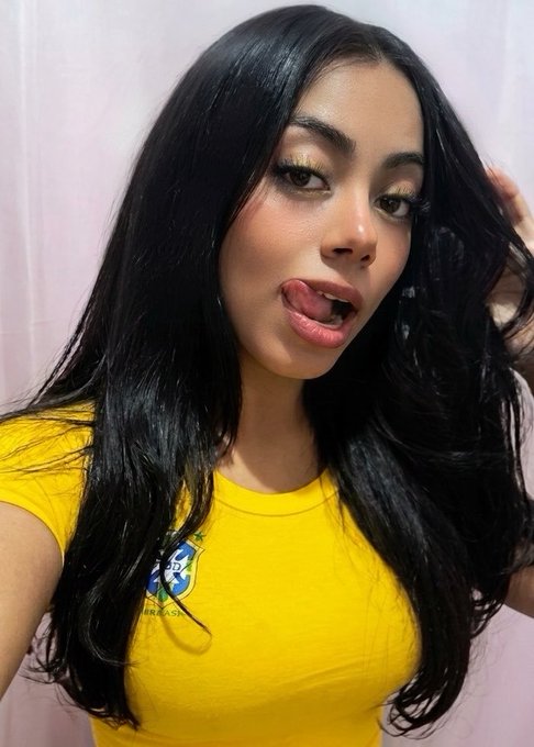 A young woman with long dark hair poses playfully with her tongue out, wearing a yellow Brazil t-shirt featuring the iconic crest. The image accompanies a promotional post encouraging viewers to help her win a 1 BTC prize, using her vibrant selfie as an engaging visual to attract support.
