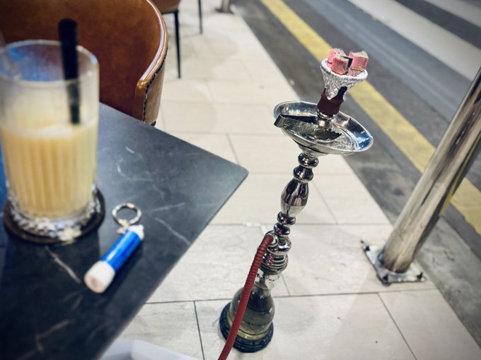 A hookah with burning coals sits next to a tall glass of a creamy beverage with a straw and a blue lighter on a dark marble table.