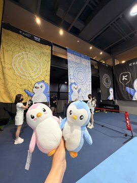 A hand holds up two penguin plush toys in front of a KRAFT LIVE event backdrop featuring similar penguin mascots.