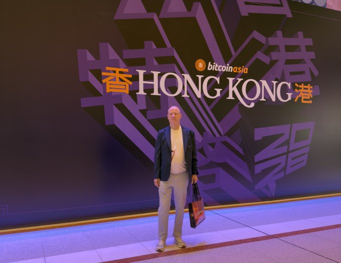 A man poses in front of a vibrant purple Bitcoin Asia "HONG KONG" backdrop featuring Chinese characters and the event's logo, capturing his attendance at the cryptocurrency conference.