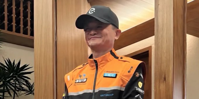 A man wearing a black cap with an 'e' logo and an orange and black team jacket with sponsor logos including 'Alibaba Cloud', 'CNBC', 'Eurodat', and 'Ant Group' smiles in an indoor setting.