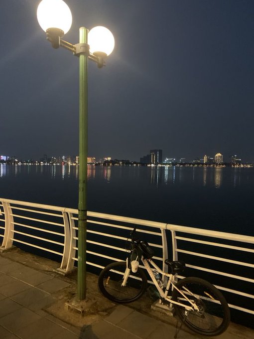 A nighttime waterfront scene with a bicycle leaning against a white railing, illuminated by a double-globe lamppost, with city lights reflecting across the water in the background.