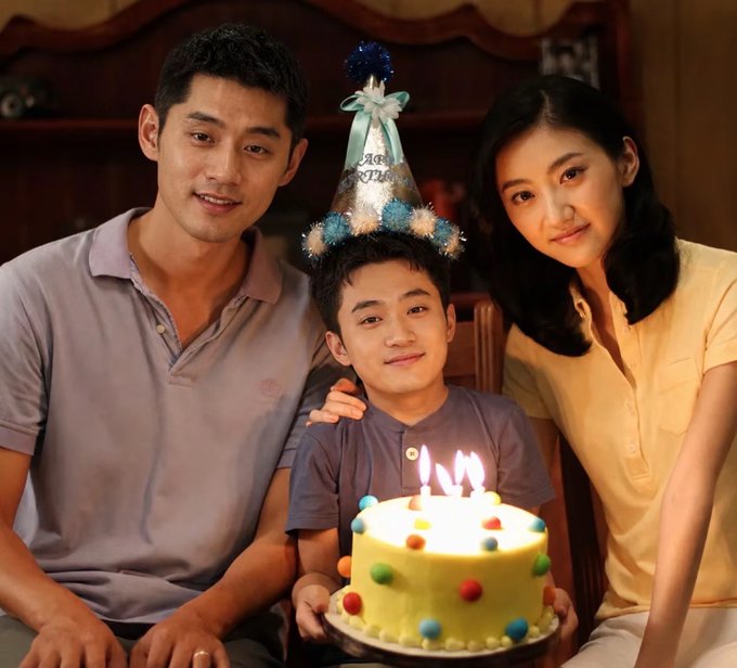 A family of three—a man, woman, and boy wearing a birthday hat—gathers around a cake with lit candles and colorful decorations, smiling warmly in a softly lit indoor setting.