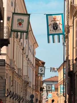 Colorful heraldic flags bearing coats of arms hang above a narrow Italian street, with one flag reading "CITTÀ DI PARMA," showcasing the historic architecture typical of a journey through Italy's Piemonte region. The scene captures the charm and traditional character that complements the trip to Southern Milan's surrounding areas.