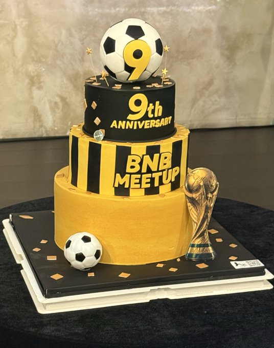 A three-tiered 9th anniversary cake for BNB Meetup, decorated in black and yellow with a soccer ball topper and a FIFA World Cup replica trophy.