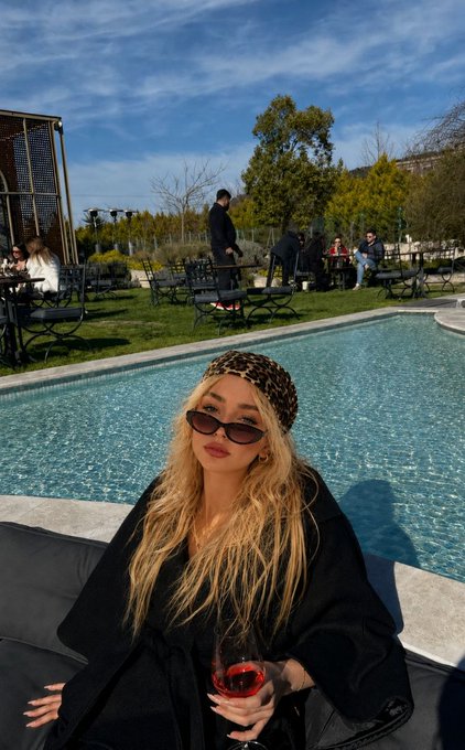 A stylish woman wearing a leopard-print headband and sunglasses lounges poolside with a glass of red wine, dressed in a black coat — the mood of this relaxed morning scene is being shared with the crypto community to ask, "Good morning crypto family! Which coin will be the most talked about today? Let's see if the first person to guess will be right?"