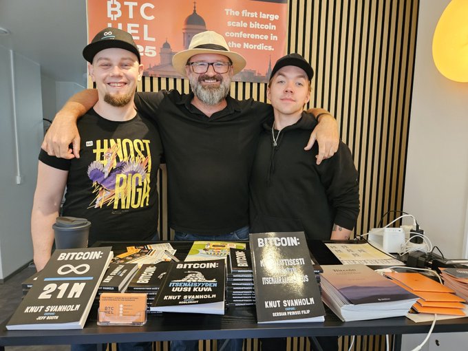 Three men pose behind a table displaying books and merchandise at a BTC Helsinki 2025 event, with the central man wearing a straw hat and the books titled 'BITCOIN: 21M' and 'BITCOIN: UUSI KUVA' by Knut Svanholm.