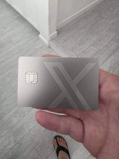 A first-person photo of a hand holding a sleek, silver metal debit card featuring an embossed 'X' logo and a smart chip, taken over a light wood floor with a foot in a black flip-flop visible below.