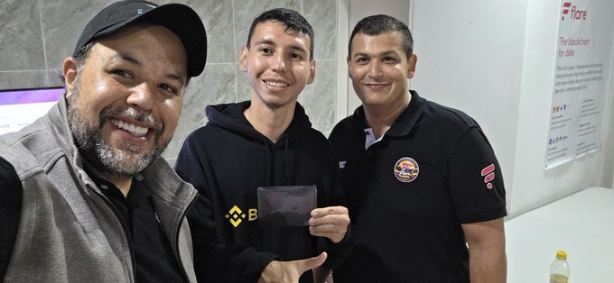 A selfie of three smiling men indoors; the middle person in a black hoodie is holding what appears to be a card or license, while the man on the right wears a 'K-Paul's' logo shirt, with a 'Bitcoin' informational poster visible on the wall behind them.