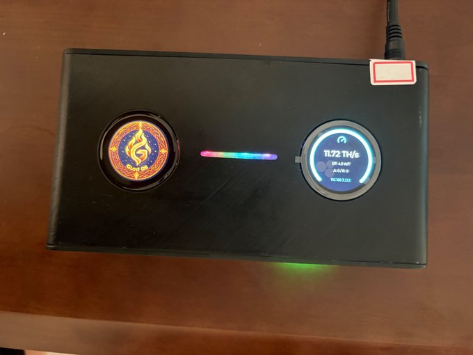 A black electronic device on a wooden surface, possibly a miner, featuring a circular logo, an RGB light strip, and a digital display reading '11.72 TH/s' with hashrate statistics.