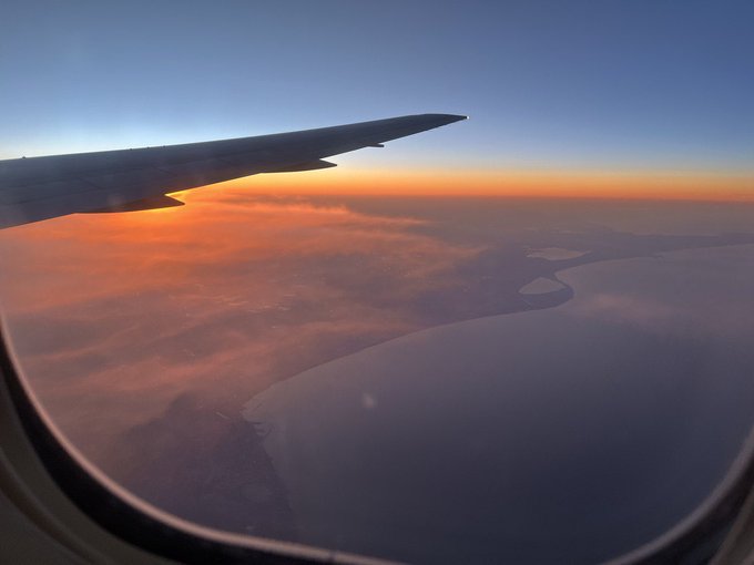 The image shows an airplane window with a view of clouds below, as well as a sunset over the horizon. The sky appears hazy due to the sun setting behind the clouds. The photo has been edited to include text that reads "New DROP Between Horizons objkt.com/tokens/KT1DzCB". This suggests that the image may have been taken during a flight and then edited for publication or sharing on social media platforms.

The airplane window provides an interesting perspective of the sky, clouds, and sunset, creating a visually appealing scene. The photo captures the beauty of nature as it transitions from day to night, with the sun setting behind the clouds and casting a warm glow over the landscape below.