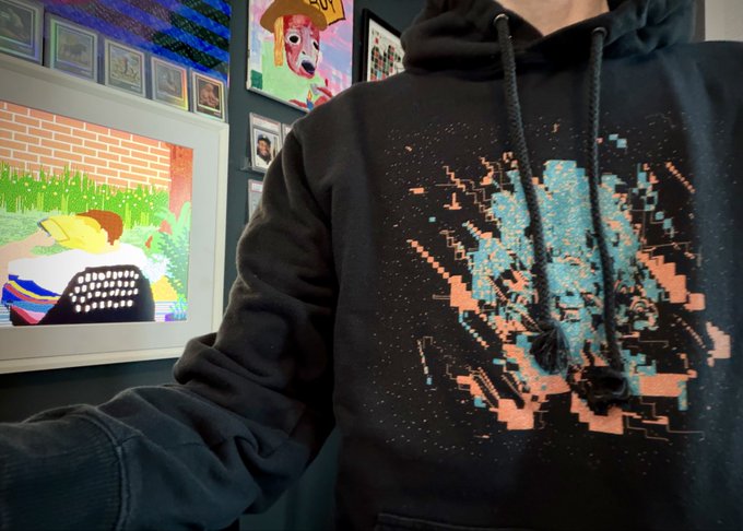 A person wears a black hoodie featuring a glitch-art design with fragmented blue and orange pixelated patterns resembling a digitally distorted figure. Behind them, a wall displays vibrant framed artworks, complementing the post's message that each "Remnants" hoodie is handmade individually, emphasizing its artisanal craftsmanship.