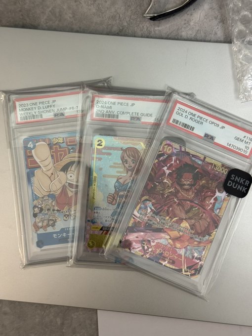 Three graded One Piece Japanese trading cards (Monkey D. Luffy Shonen Jump, Nami Complete Guide, and Gol D. Roger) sit in PSA slabs, with a "SNKR DUNK" NFT sticker—a perfect intersection of the poster's love for TCGs and NFTs.