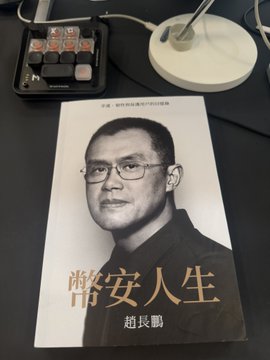 A hand-signed copy of "幣安人生" (Binance Life) by Changpeng Zhao (CZ) rests on a desk next to a Stream Deck, with CZ's signature visible above his printed portrait on the cover. The book, a gift from the Binance founder, features his black-and-white photo and Chinese title on the front.