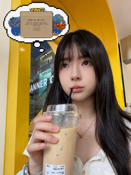 The image features a woman with long black hair holding a drink, possibly an iced coffee or tea, while looking at the camera. The background includes a yellow door and a window, suggesting that she might be inside a store or cafe. There is also a computer screen visible in the scene, which could indicate that this location is part of a business or workspace.