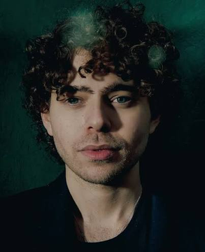 The image features a man with curly hair, dressed in black clothing, posing for a picture against a green background that gives off a grunge vibe. The photo suggests an underground or alternative lifestyle scene, as one of the statements in the image mentions the Polymarket website where fake bets were being staged by creators. This information implies that the subject could be part of some kind of online community or event related to gambling and alternative lifestyles.