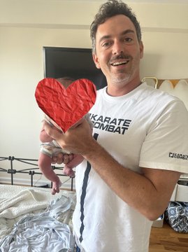 A smiling man with a mustache wearing a "Karate Combat" t-shirt holds a red heart-shaped object, while cradling a baby in his other arm. The post celebrates love and tags @tickerBITCOINbb, with the heart symbol reinforcing the affectionate message of "We love you!!!"