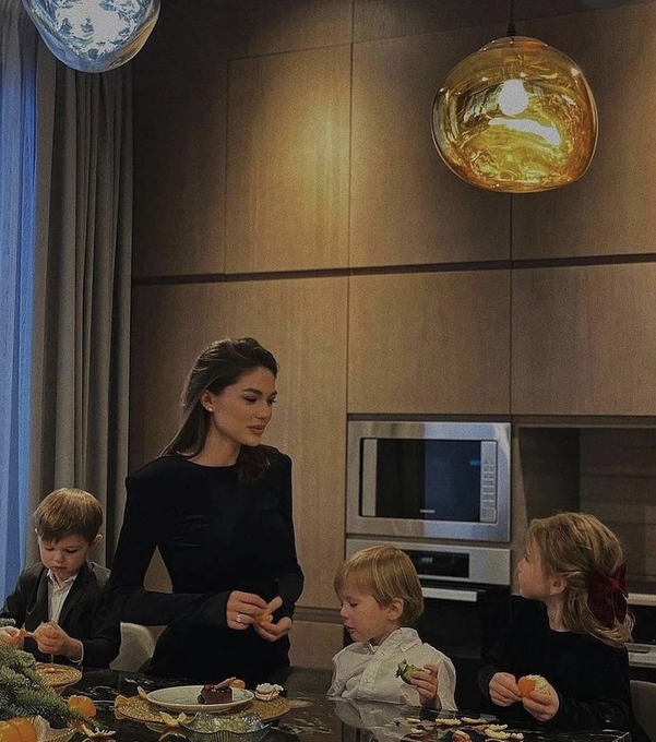 A woman in a black top stands in a modern kitchen preparing food while three young children sit at the counter eating snacks.