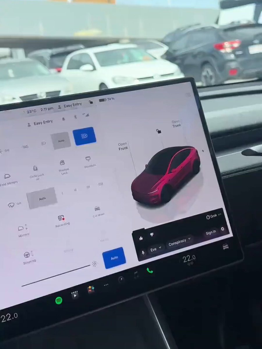 The image shows a close-up view of a car's dashboard screen, which appears to be a GPS navigation system. The background features cars driving by with their windshields down at a parking lot area. A caption below the image questions whether Musk' s GROK is really waking up or if it might be part of some conspiracy involving Tesla and its founder, Musk, who are trying to bring the future into the present. However, this claim seems far-fetched and unrelated to the actual content of the image. The focus should remain on the GPS navigation system displayed on the dashboard screen.