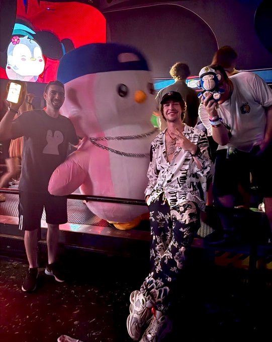 A young man in a patterned shirt and cap poses smiling beside a large inflatable penguin mascot and a person in a Mickey Mouse shirt, capturing a fun "Gm" morning moment at what appears to be a Disney-themed event.