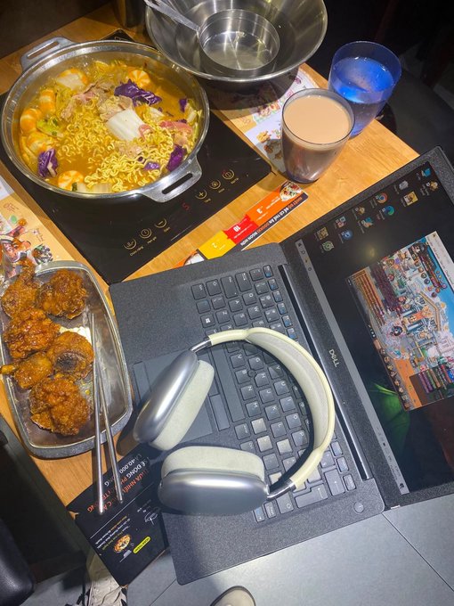 A workspace scene showing a laptop with headphones resting on the keyboard, alongside a hot pot with instant noodles, a dish of fried food, and beverages on a wooden table.