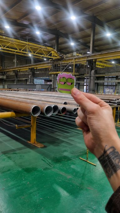 A hand with a tattoo holds up a small green square charm featuring a cartoon face in an industrial setting with large metal pipes.