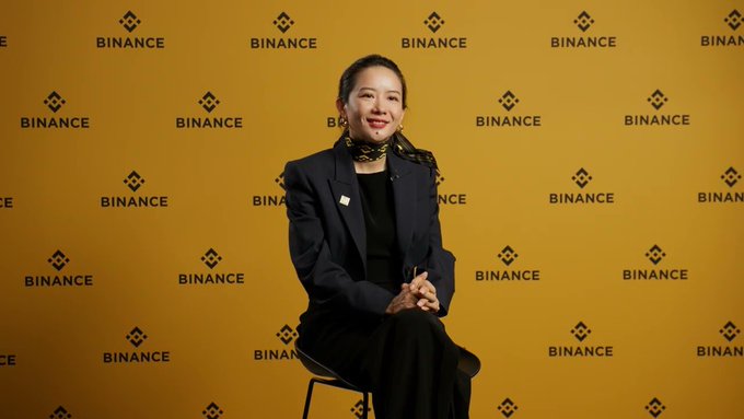 The image features a woman sitting on a chair, wearing a black jacket and pants. She has her hands together and appears to be smiling at the camera. The background of the image consists of yellow text that reads "BINANCE" and "ta ce". This suggests that she is in an office setting or a room with a company logo theme.