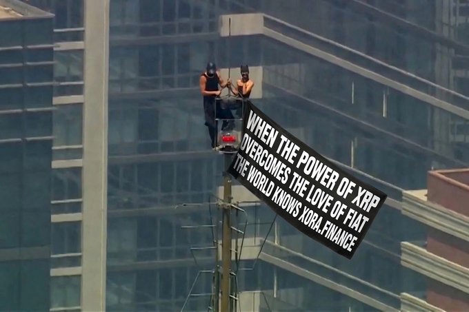 Two shirtless protesters stand atop a tall structure in an urban setting, unfurling a black banner reading "WHEN THE POWER OF XRP OVERCOMES THE LOVE OF FIAT THE WORLD KNOWS XORA.FINANCE," echoing the post's emphasis on XRP's potential to surpass traditional fiat currency systems.