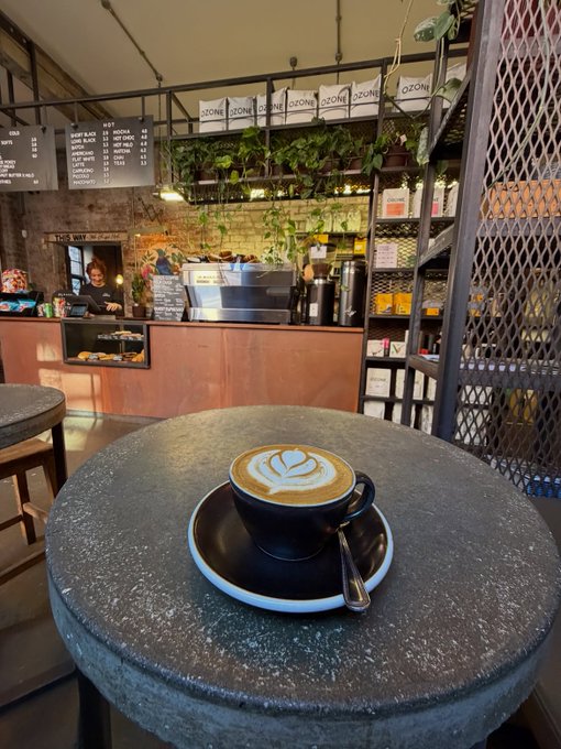A cappuccino with latte art served in a black cup on a stone table at an Ozone coffee shop, with the cafe's counter, menu boards, and a barista visible in the background.