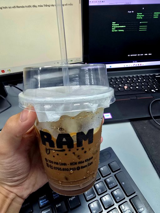 A hand holding a clear plastic cup with a domed lid and straw containing an iced coffee, labeled 'RAM', held in front of a laptop keyboard and screen.