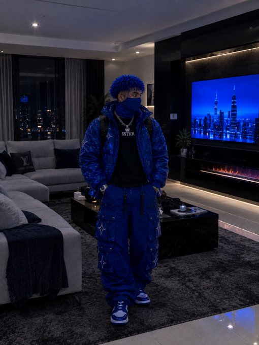 A person stands in a modern living room wearing an all-blue puffer jacket, cargo pants, and a matching blue mask and curly wig, with 'STTKR' visible on their shirt.