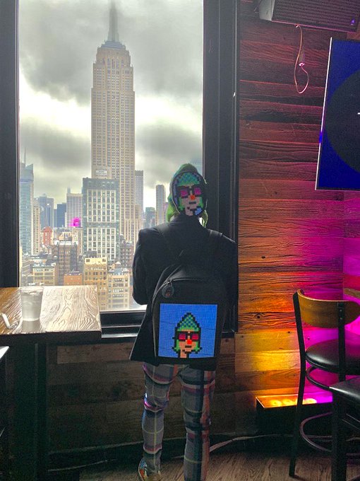 A person wearing a pixel-art mask and a matching backpack stands by a window overlooking the Empire State Building in New York City.