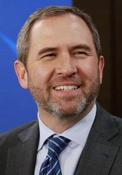 A smiling man with a beard wearing a blue and white striped tie and dark suit stands in front of a blue background. His cheerful expression contrasts sharply with the post announcing that $XRP has plunged 13%, suggesting an ironic disconnect between his positive demeanor and the negative crypto market news.