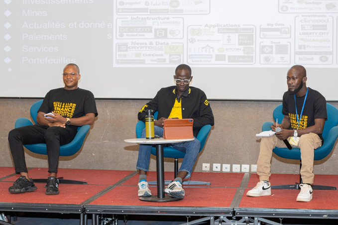 Three men in yellow and black 'WILL BE FINANCE SURPRISE' shirts sit on blue chairs on a stage with a presentation about fintech topics like investments, mining, payments, and services displayed behind them.